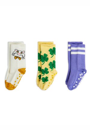 Tre par strumpor: vita med kattdesign och gula accenter, gula med gröna klöversmönster och lila med vita ränder och grepp.