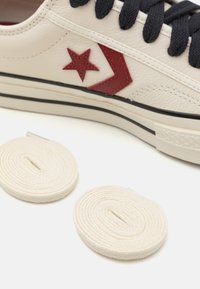 Gros plan sur une basket beige avec des lacets noirs et un logo étoile rouge, présentant deux lacets blancs roulés sur une surface blanche.