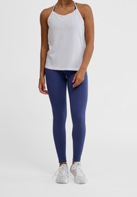 Canotta atletica bianca con design a halter e logo; abbinata a leggings blu scuro a coste e sneakers grigio chiaro.