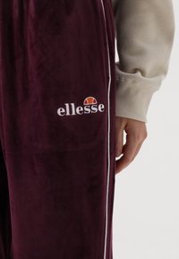 Vinsorte velour tracksuit-bukser med hvide sidelister og et broderet "ellesse"-logo med et rødt og orange emblem.