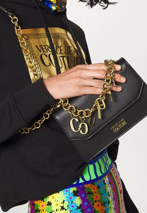 Person med en svart Versace Jeans Couture huvtröja och en färgglad blommig kjol, som håller en svart läderväska med guldkedja och logocharmar.