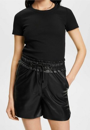 Ung person iført en sort, ribstrikket kortærmet skjorte og højtaljede sorte faux lædershorts med snøre i taljen.