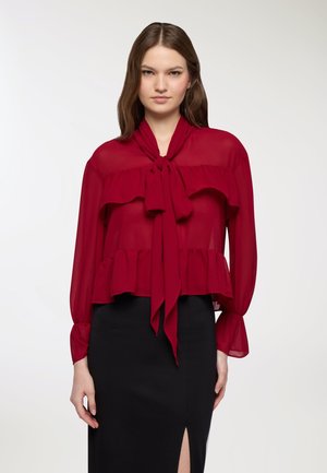 Blusa rossa trasparente con colletto a fiocco e orlo a strati. Maniche lunghe con polsini abbottonati. Abbinata a una gonna nera aderente.