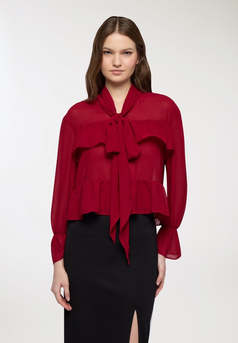 Blusa rossa trasparente con colletto a fiocco e orlo a strati. Maniche lunghe con polsini abbottonati. Abbinata a una gonna nera aderente.