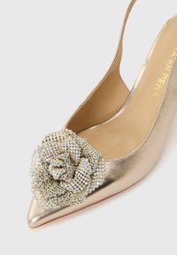 Χρυσό μυτερό γοβάκι ψηλού τακουνιού με ένα μεγάλο λαμπερό λουλούδι από στρας στη μύτη.