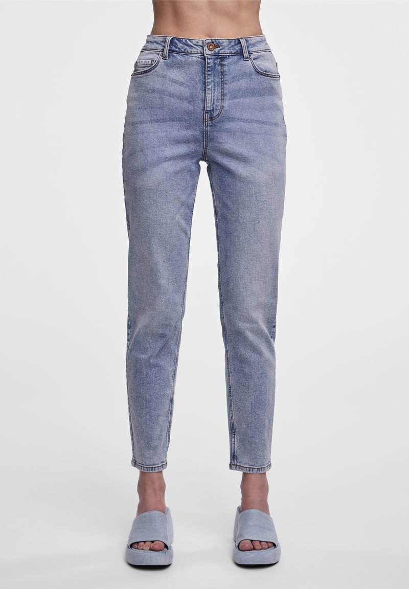 Femme portant un jean denim bleu clair taille haute et des sandales à plateforme bleu clair assorties à bouts ouverts, debout devant un fond blanc.