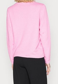 Rosa strukturierter Pullover mit einer reliefartigen Strickmusterung, langen Ärmeln, rundem Ausschnitt und geripptem Saum, kombiniert mit schwarzen Hosen.