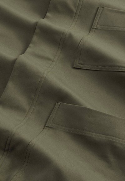 Tkanina oliwkowa o gładkiej fakturze, widocznych szwach i wielu kieszeniach naszywanych w ujęciu z bliska, podkreślająca trwałość i design.