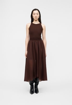 Kvinde iført en brun, ærmeløs kjole med højtaljet pasform og sorte knælange støvler, stående mod en ensfarvet lys baggrund.