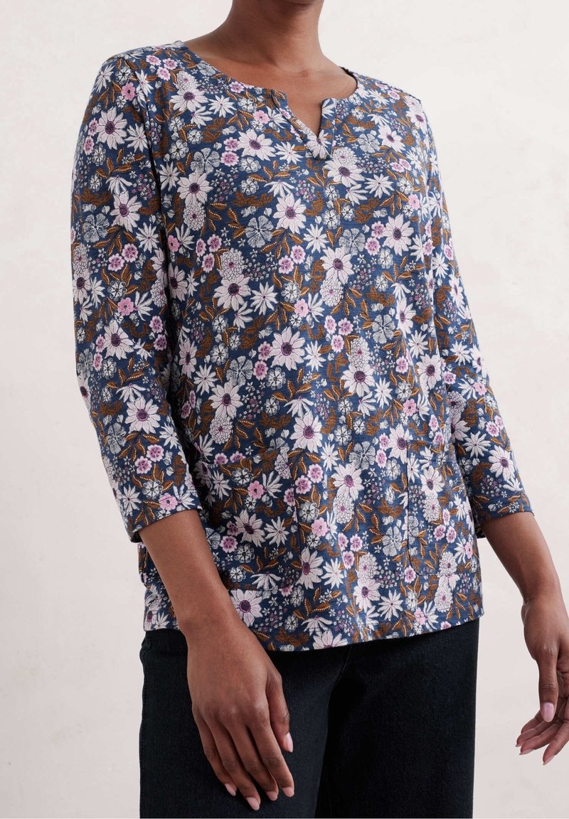 Kvinna i en långärmad blå blommig topp med rosa, vita och bruna blommor, kombinerad med svarta byxor mot en enkel bakgrund.