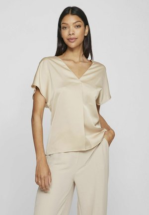 Haut en satin beige avec un col en V et des manches courtes. Présente une coupe ample et un drapé subtil, associé à un pantalon beige. Texture lisse.