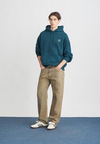 Hoodie teal avec une poche avant et un logo, associé à un jean droit beige clair et des baskets blanches avec des rayures noires.