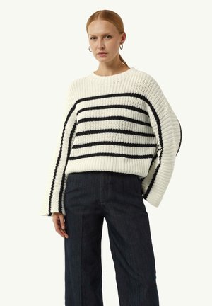 Chunky gebreide trui in crème met zwarte horizontale strepen, met brede mouwen en een relaxte fit. Gecombineerd met donkere broeken.
