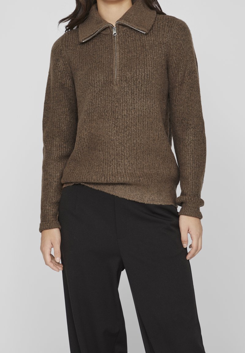 Femme portant un pull en maille marron avec un col zippé au quart et un pantalon noir, debout devant un fond clair uni.