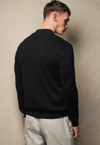 Černá pletená mikina s kulatým výstřihem, žebrovanými manžetami a lemem, nošena s světle šedými kalhotami, která ukazuje minimalistický design.