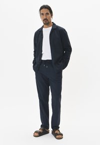 Camisa de botones azul marino con bolsillo en el pecho, camiseta blanca, pantalones azul marino con cordón, y sandalias negras con detalles de hebilla, de pie sobre un fondo liso.