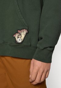 Πράσινο φούτερ με μπροστινή τσέπη καγκουρό, διακοσμημένο με πολύχρωμο κεντημένο σχέδιο μανιταριού στο κάτω αριστερό μέρος. Υφασμα από μείγμα βαμβακιού.