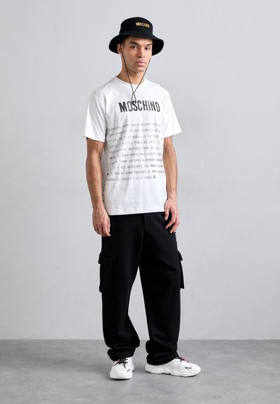 Hvit bomull t-skjorte med "MOSCHINO" trykket i svart, kombinert med svarte cargo-bukser og hvite sneakers. Tilbehøret inkluderer en svart bøttehatt.