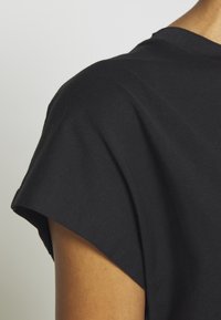 Zbliżenie na ramię osoby noszącej czarną bawełnianą koszulkę z krótkim rękawem na jednolitym jasnoszarym tle.