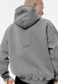 Persona che indossa una felpa grigia oversized con cappuccio alzato e un piccolo logo ricamato sulla schiena, sfondo chiaro.