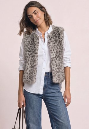 Femme portant une chemise blanche boutonnée, un gilet en fourrure gris à taches, un jean bleu et tenant un sac à main marron sur un fond clair uni.
