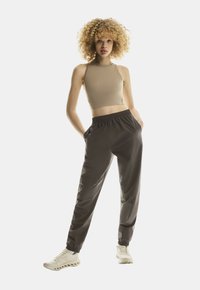 Mlada osoba s kovrčavom kosom, odjevena u bež bezrukavni crop top, tamnosive trenirke i bijele tenisice, stoji s rukama u džepovima.