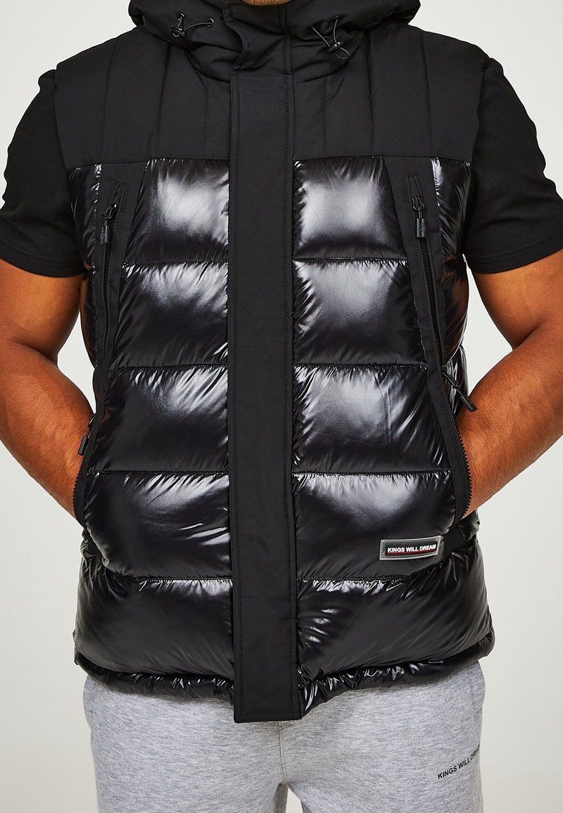 Gilet rembourré noir avec finition brillante, capuche et poches zippées. Présente un mélange de matériaux mats et brillants, avec une étiquette logo en bas.
