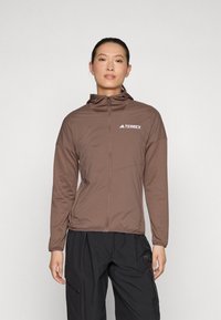 Giacca marrone con zip e cappuccio, realizzata in materiale leggero e testurizzato. Presenta il logo "Terrex" sul lato superiore destro.
