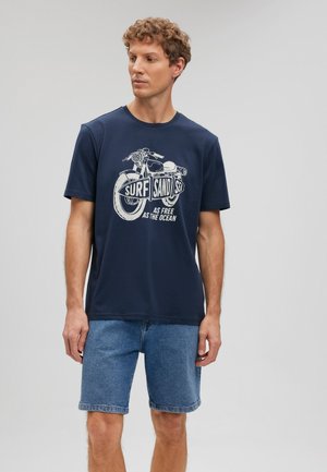 Navy blue cotton t-shirt featuring a graphic of a motorcycle with text "SURF SAND SEA AS FREE AS THE OCEAN." Paired with denim shorts.