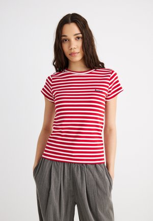 Giovane donna con capelli lunghi che indossa una maglietta a righe orizzontali rosse e bianche e pantaloni grigi a righe sottili, con le mani nelle tasche.