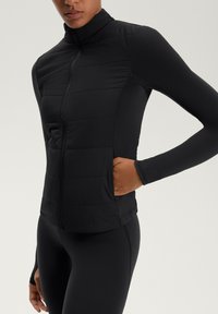 Donna che indossa una giacca nera attillata trapuntata con maniche lunghe e zip frontale, con una mano nella tasca laterale, su uno sfondo leggero uniforme.