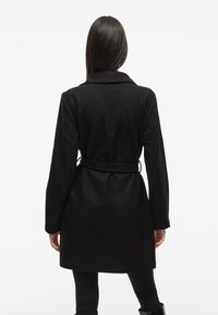 Abrigo de lana negro con cintura ceñida por cinturón, solapas con muesca y mangas largas; textura suave y diseño hasta la rodilla; silueta minimalista y clásica.