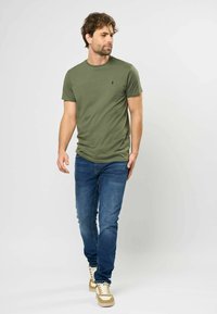 Olivinės spalvos trumpų rankovių marškinėliai su apvaliu kaklu ir mažu logotipu, derinami su mėlynomis džinsomis ir šviesiai rudais sportbačiais. Lygus, kasdieniškas siluetas.