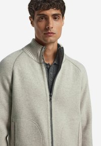 Hellgrauer Zip-Hoodie mit geripptem Kragen, ausgestattet mit Fronttaschen und einer Schicht aus dunkelgrauem Stoff darunter. Weiche Textur und taillierte Passform.