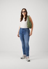 Modre kavbojke slim fit, bela majica brez rokavov, zelena torbica preko telesa in sivi športni čevlji. Dodatki vključujejo pravokotne sončne očala.
