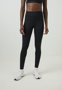 Fekete leggings magas derekú, sima textúrával, testhezálló kialakítással és látható minták nélkül. Fehér sportcipőkkel viselve.