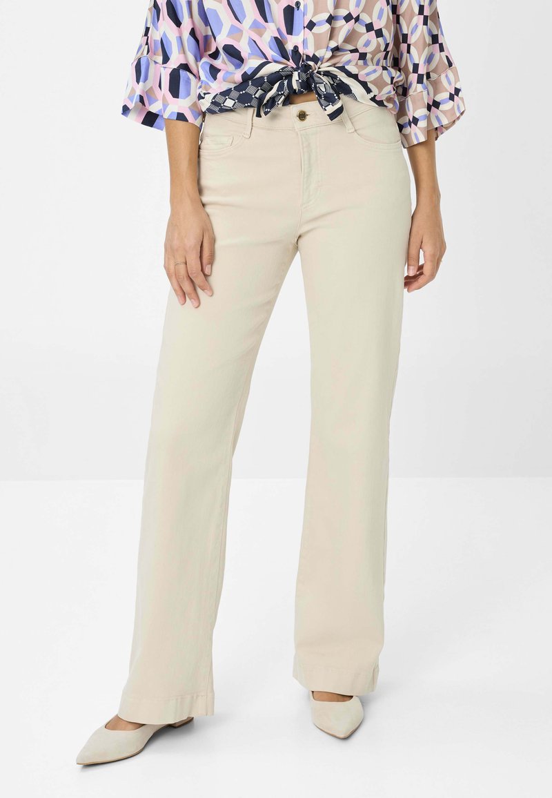 Beige wijde broek met voorzakken en een lichte glans, gecombineerd met een gebloemd blousje. De schoenen zijn lichtgekleurde pumps met een spitse neus.