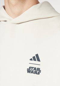 Lys beige hættetrøje lavet af blødt stof med et mørkeblåt Adidas-logo og "STAR WARS" tekst på brystområdet.