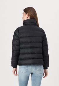 Svart dunjakke med quiltede seksjoner, høy krage og ribbestrikkede mansjetter, paret med lysvaskede jeans. Myk, blank ytre finish.