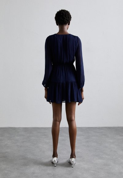 Rochie mini pliată bleumarin, cu mâneci lungi și talie elastică, având o fustă evazată și tocuri argintii cu vârf ascuțit.