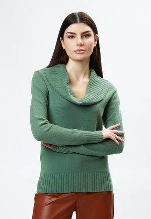 Maglione verde con collo a cowl, texture a coste sul colletto e sui polsini, e un corpo aderente. Il tessuto appare morbido e caldo.