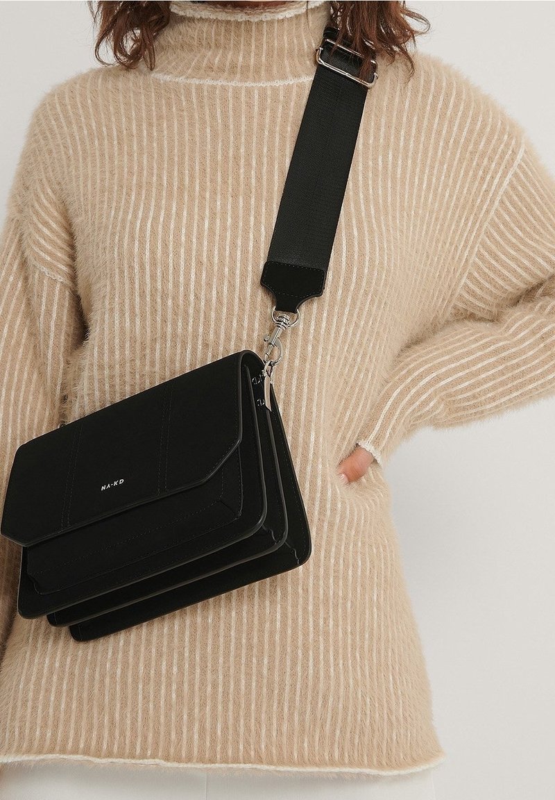 Musta olkahihnalla varustettu laukku, jossa on kolme päällekkäistä lokeroa, sileä pinta ja hopeiset yksityiskohdat. Laukku on päällä beige ribbipaitaan.