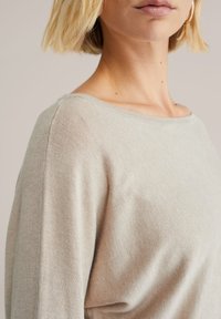 Gaiši bēša adīta džemperis ar plašu kakla izgriezumu un ribotu tekstūru. Tam ir maiga, gluda apdare un atbrīvota piegrieztne ap pleciem.