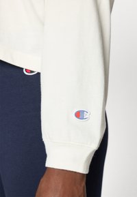 Kremfarget langermet skjorte med ribbede mansjetter, som har en blå og rød brodert logo på ermene. Marineblå bukser delvis synlige nedenfor.