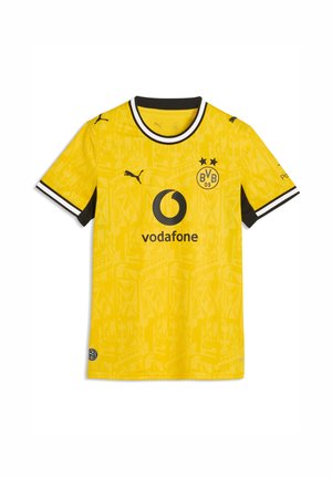 Mujer con camiseta amarilla del Borussia Dortmund sosteniendo un balón de fútbol negro y amarillo a juego, de pie frente a un fondo gris.