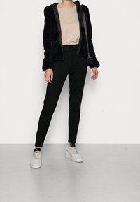 Giacca nera pelosa con zip sopra una maglietta beige a maniche corte, abbinata a pantaloni neri aderenti e sneakers bianche, con un design elegante.