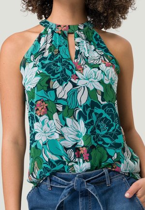 Femme portant un haut sans manches à col nu avec un motif floral turquoise, vert, blanc et rose, associé à un pantalon en jean bleu à taille nouée.