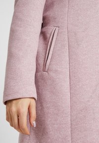 Roze wollen jas van een wollenmix met een gestructureerde stof, voorzien van een zijzak en een getailleerde silhoët, weergegeven op een hand.