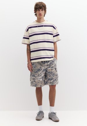 Mladý muž v krémovém a fialovém pruhovaném oversized tričku, vzorovaných cargo šortkách, bílých ponožkách a šedých teniskách, stojí s jednou rukou v kapse.