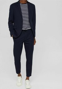 Granatowa marynarka o dopasowanym kroju w połączeniu z t-shirtem w paski, pasującymi spodniami i białymi trampkami. Posiada jedną kieszeń na piersi oraz elegancki design.